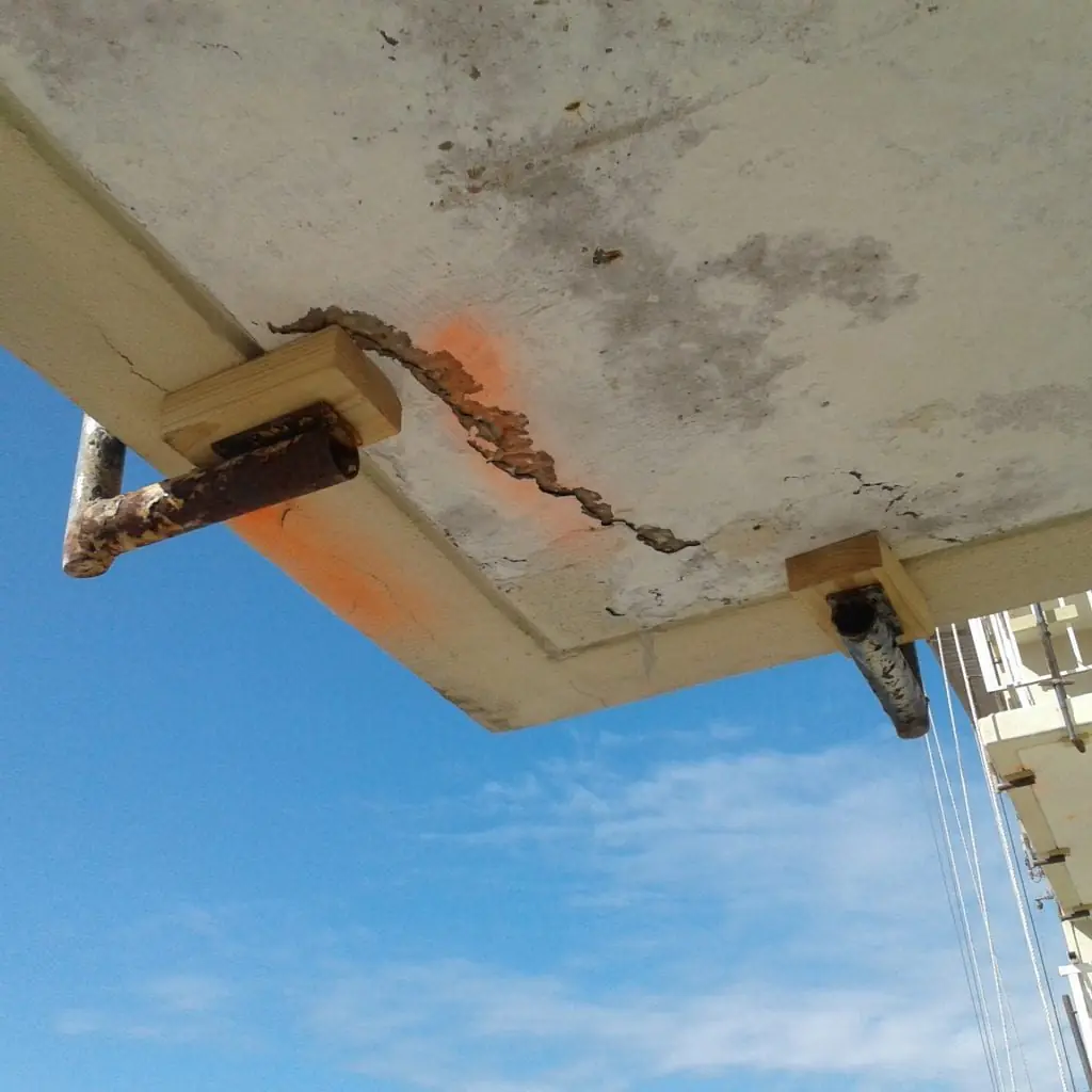 Concrete spall at a balcony edge with exposed corroded reinforcing steel, viewed from below against the sky, Fort Walton Beach, Florida.